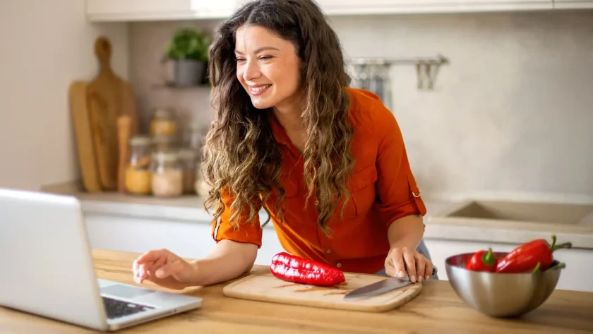 Eine Frau mit orangener Bluse steht in der Küche und guckt auf einen Laptop. Vor ihr liegt eine Spitzpaprika auf einem Schneidebrett. | VIACTIV Krankenkasse