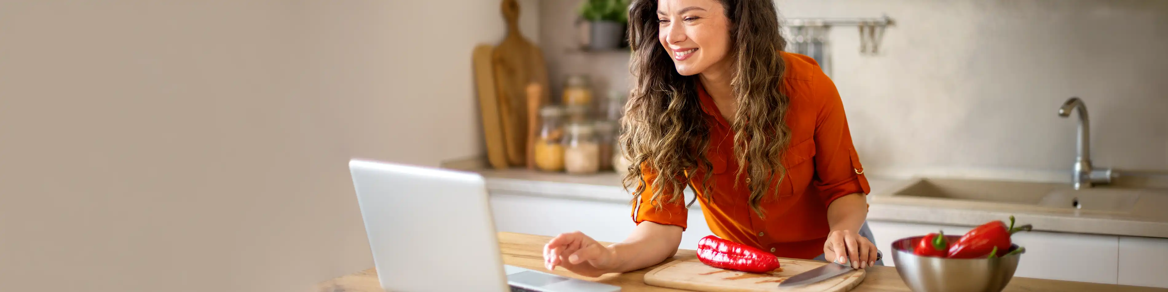 Eine Frau mit orangener Bluse steht in der Küche und guckt auf einen Laptop. Vor ihr liegt eine Spitzpaprika auf einem Schneidebrett. | VIACTIV Krankenkasse
