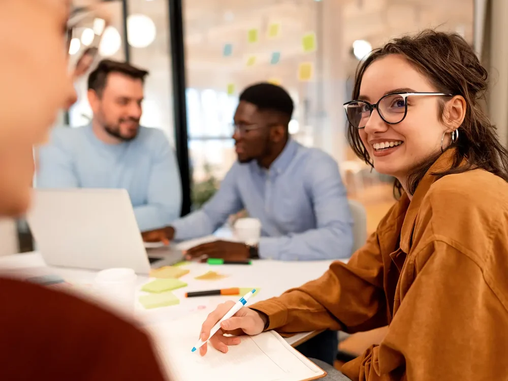 Eine junge Frau sitzt am Tisch. Sie hält einen Stift in der Hand. Vor ihr liegt ein Notizbuch. Im Hintergrund unterhalten sich zwei Männer. Vor ihnen steht ein Laptop. | VIACTIV Krankenkasse