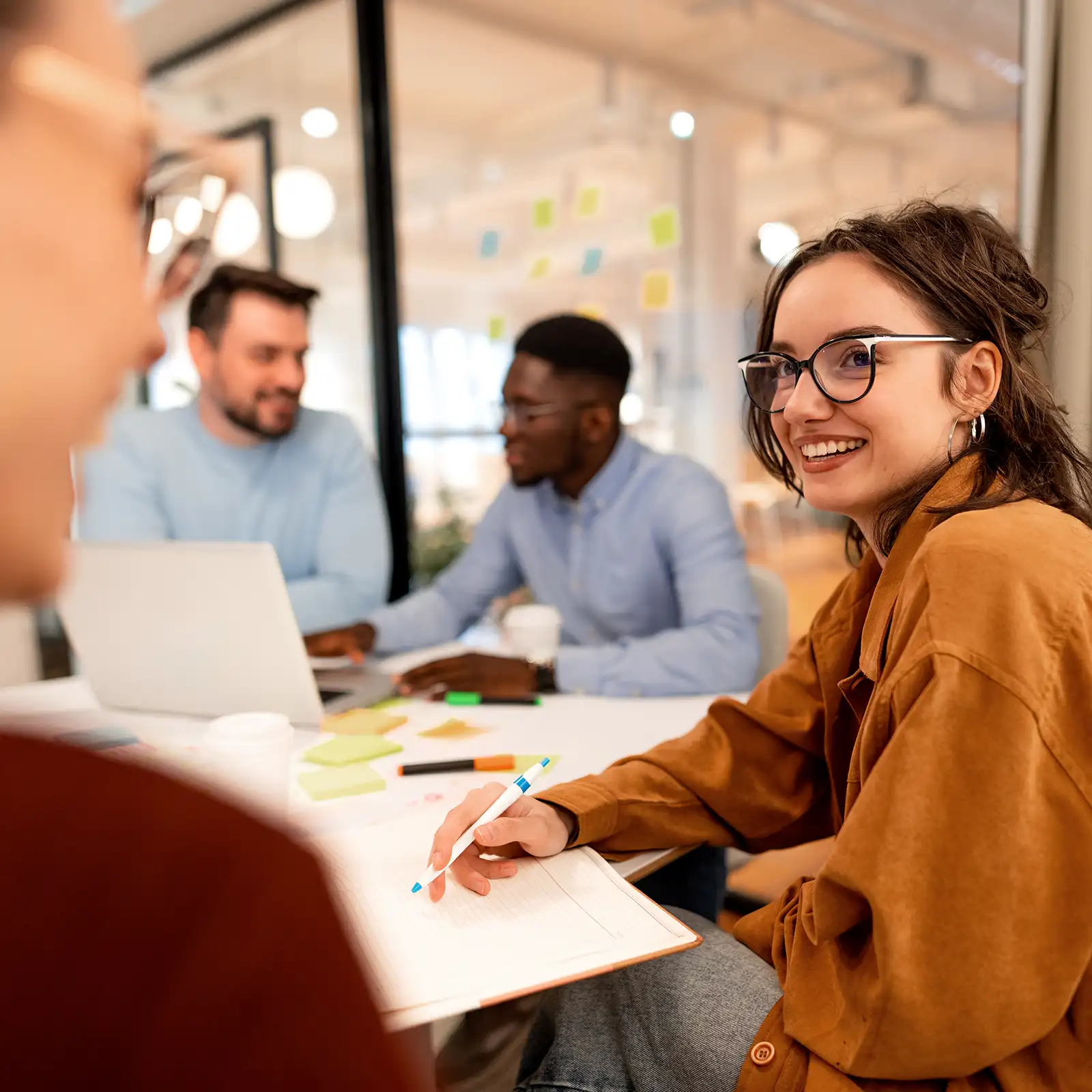 Eine junge Frau sitzt am Tisch. Sie hält einen Stift in der Hand. Vor ihr liegt ein Notizbuch. Im Hintergrund unterhalten sich zwei Männer. Vor ihnen steht ein Laptop. | VIACTIV Krankenkasse