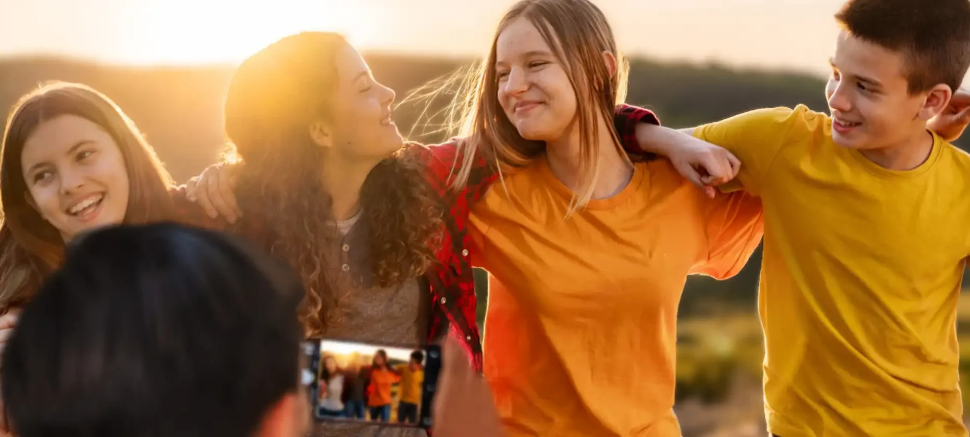 Eine Gruppe Jugendlicher hat die Arme umeinander gelegt. Sie sind in der Natur unterwegs. Vor ihnen steht ein weiterer Junge, der sie mit einem Handy fotografiert. | VIACTIV Krankenkasse