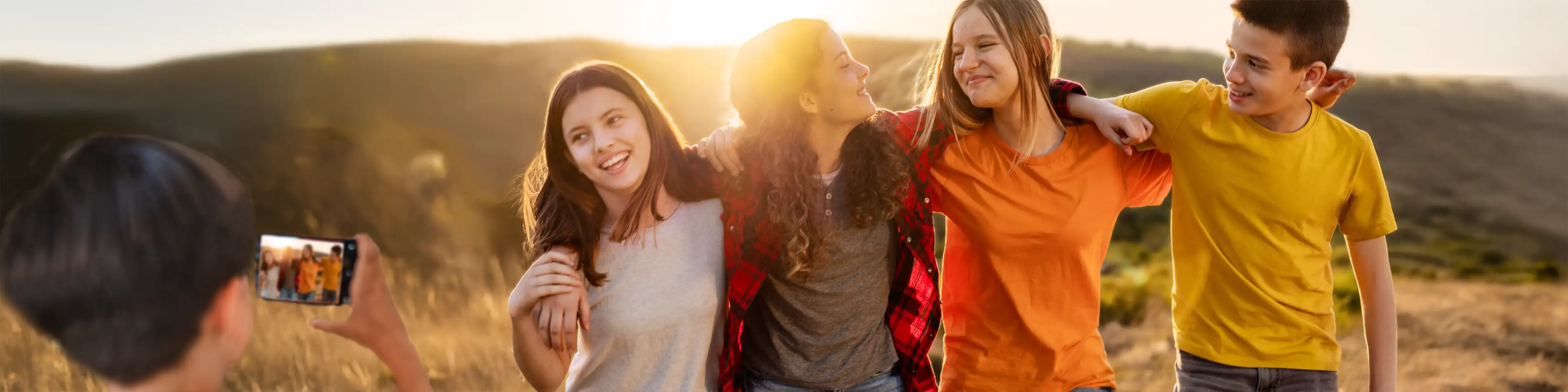 Eine Gruppe Jugendlicher hat die Arme umeinander gelegt. Sie sind in der Natur unterwegs. Vor ihnen steht ein weiterer Junge, der sie mit einem Handy fotografiert. | VIACTIV Krankenkasse
