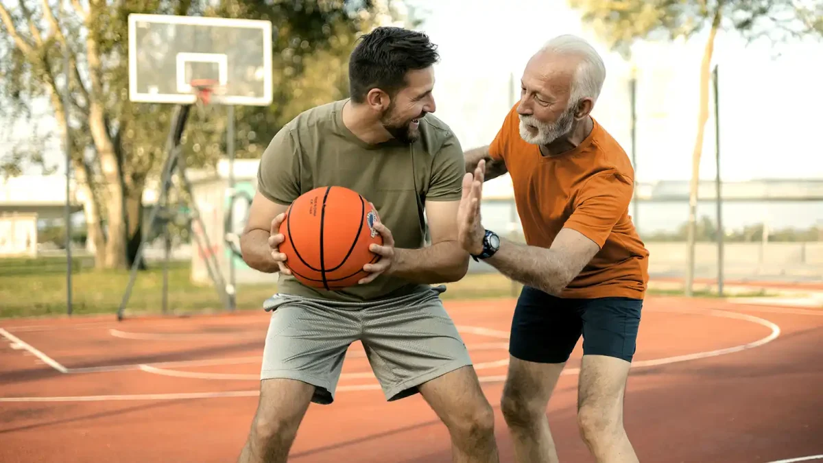 Ein jüngerer und ein älterer Mann spielen gemeinsam Basketball. Im Hintergrund ist ein Basketballkorb zu sehen. | VIACTIV Krankenkasse