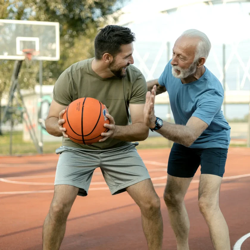 Zwei Männer spielen Basketball. Einer von ihnen ist jung, der andere etwas älter. Der junge Mann hält den Basketball in beiden Händen. | VIACTIV Krankenkasse