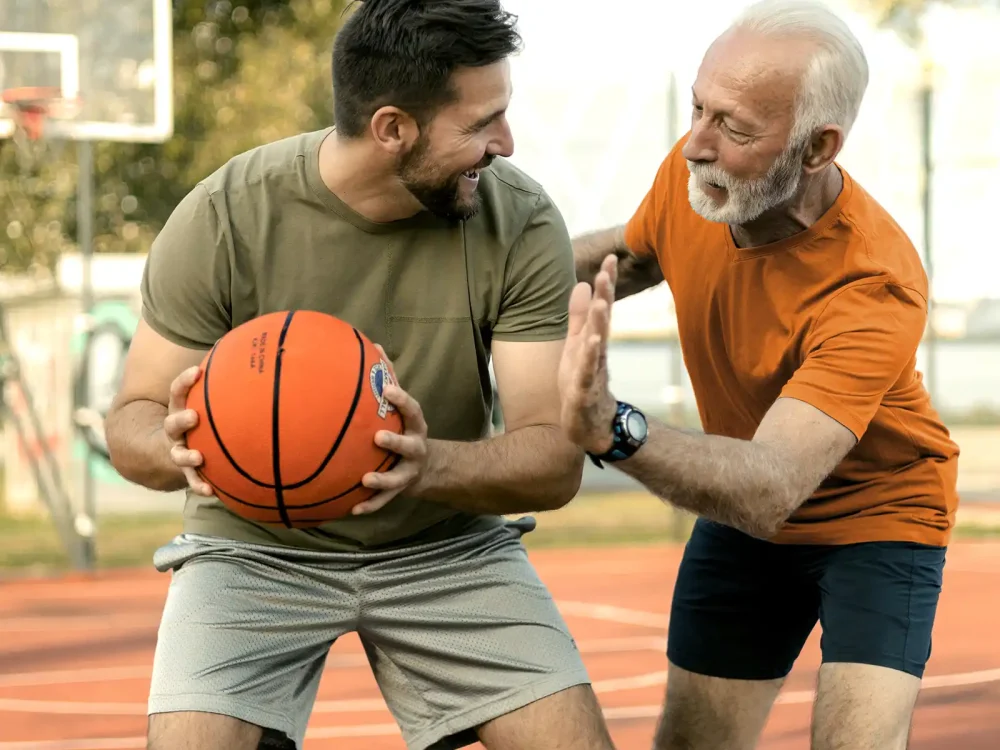 Ein jüngerer und ein älterer Mann spielen gemeinsam Basketball. Im Hintergrund ist ein Basketballkorb zu sehen. | VIACTIV Krankenkasse