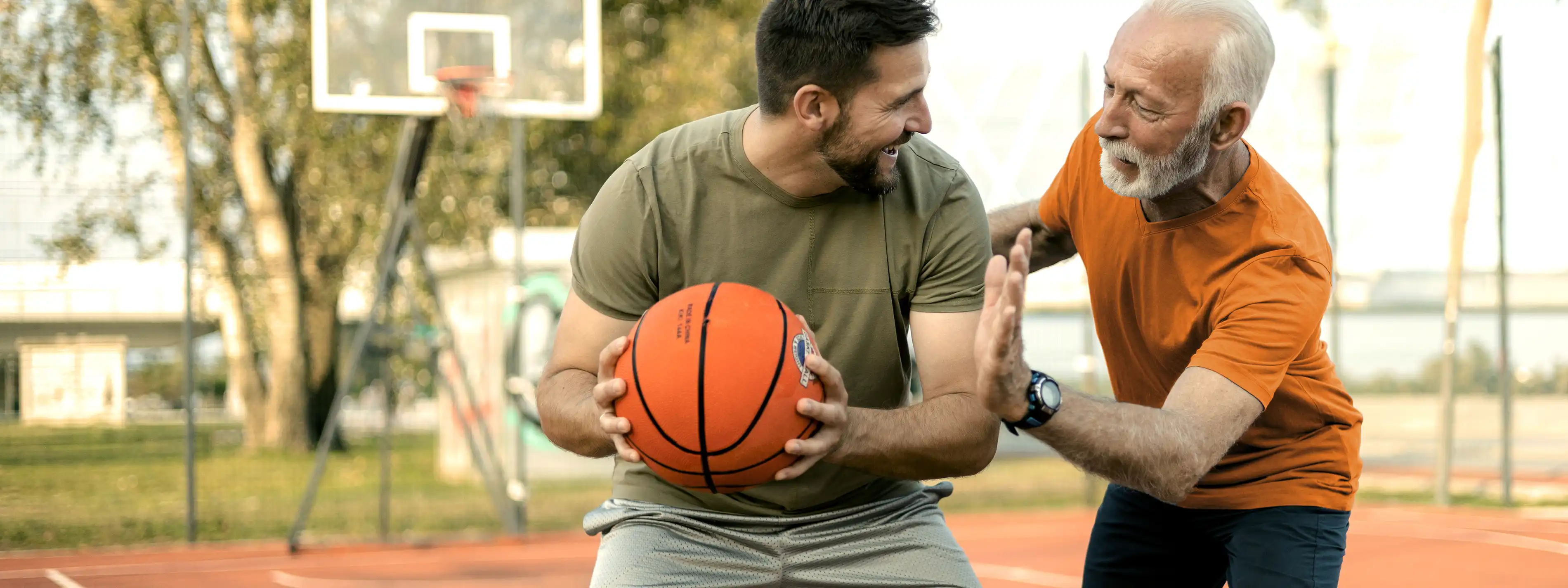 Ein jüngerer und ein älterer Mann spielen gemeinsam Basketball. Im Hintergrund ist ein Basketballkorb zu sehen. | VIACTIV Krankenkasse