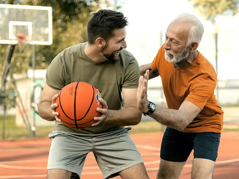 Ein jüngerer und ein älterer Mann spielen gemeinsam Basketball. Im Hintergrund ist ein Basketballkorb zu sehen. | VIACTIV Krankenkasse