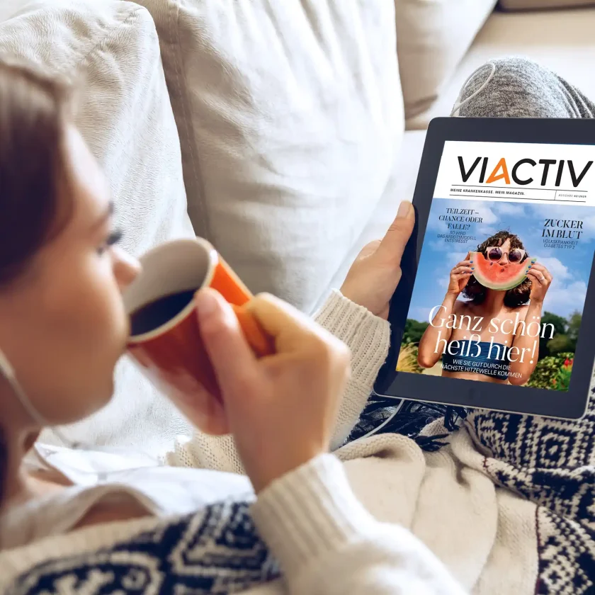 Eine junge Frau sitzt auf der Couch und liest das VIACTIV Sommermagazin auf einem Tablet. Sie trinkt dabei einen Kaffee. | VIACTIV Krankenkasse