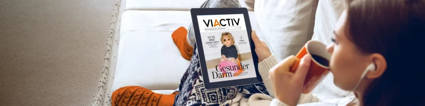 Eine Frau sitzt auf dem Sofa, trinkt Kaffee und liest auf ihre E-Book-Reader das Magazin der VIACTIV Krankenkasse. | VIACTIV Krankenkasse