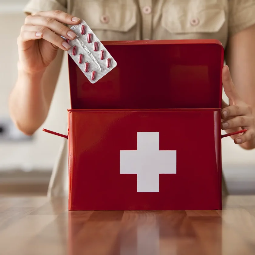 Im Vordergrund ist ein Medikamentenkoffer zu sehen. Im Hintergrund steht eine Frau, die gerade eine Tablettenpackung in den Koffer packt. | VIACTIV Krankenkasse