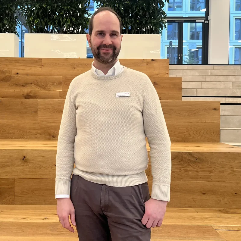 Ein Mitarbeiter der VIACTIV steht vor einer Treppe und lächelt. | VIACTIV Krankenkasse