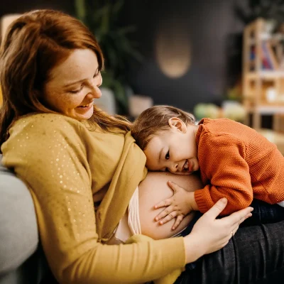 Eine junge Frau sitzt auf dem Sofa und ein kleines Kind umarmt ihren Babybauch. | VIACTIV Krankenkasse