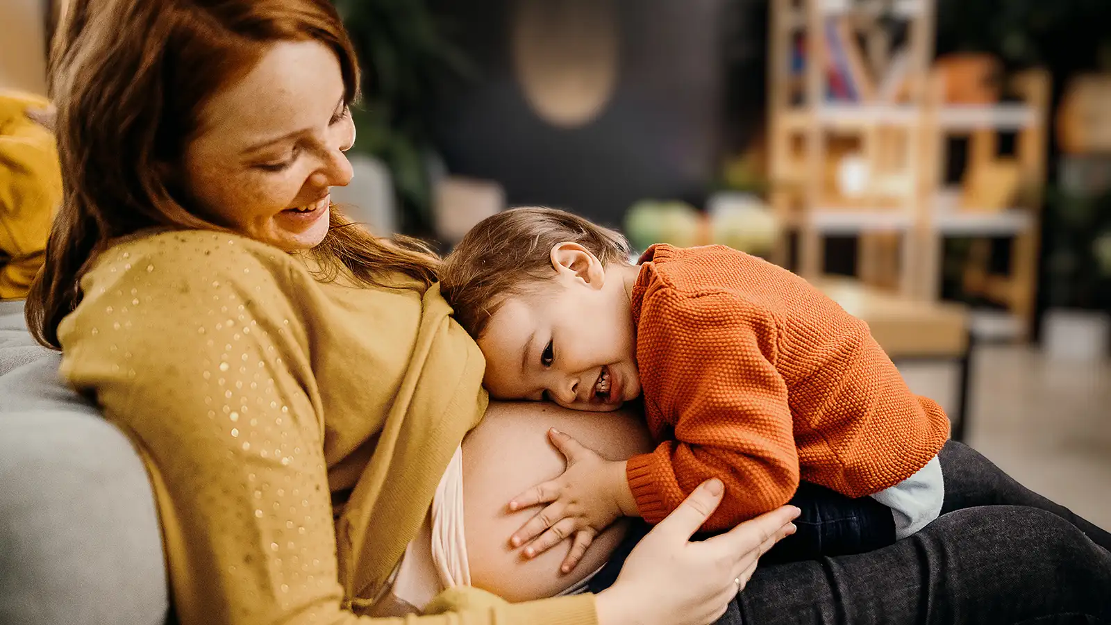 Eine junge Frau sitzt auf dem Sofa und ein kleines Kind umarmt ihren Babybauch. | VIACTIV Krankenkasse