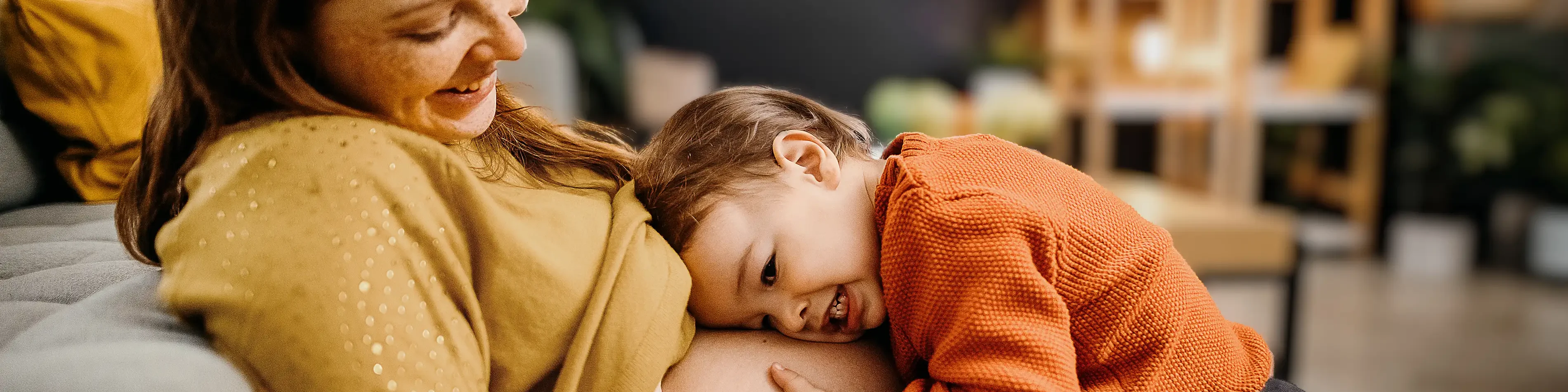 Eine junge Frau sitzt auf dem Sofa und ein kleines Kind umarmt ihren Babybauch. | VIACTIV Krankenkasse