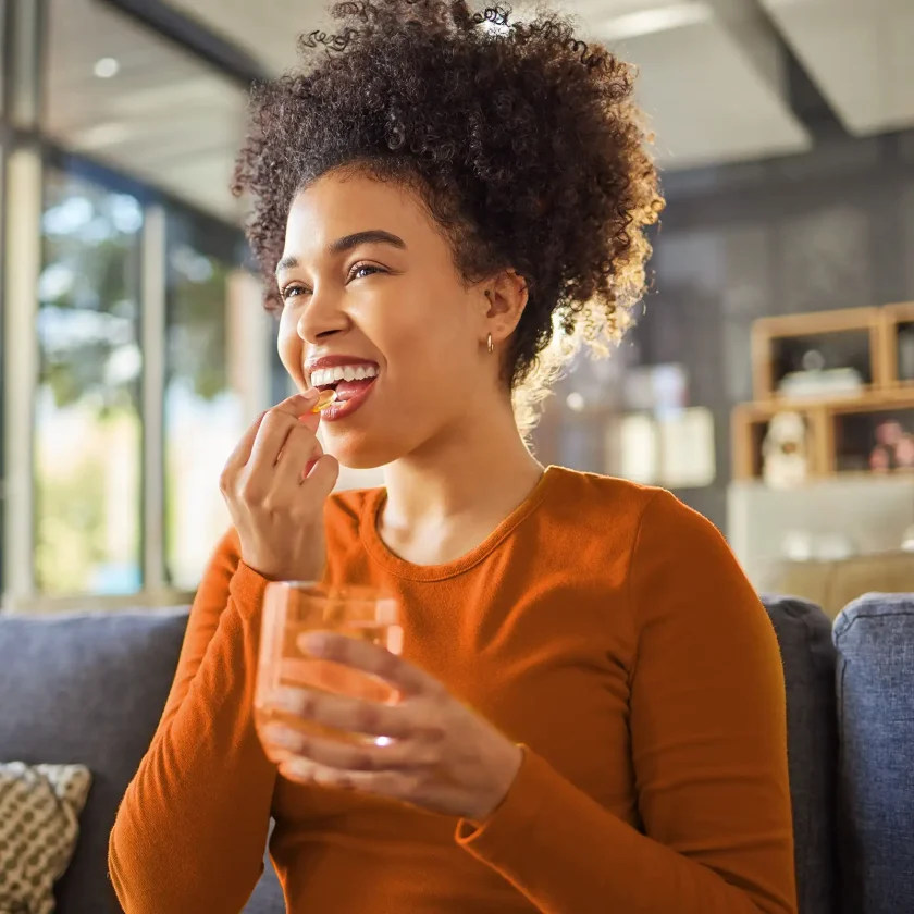 Eine Frau mit orangenem Oberteil nimmt Nahrungsergänzungsmittel. In der einen Hand hat sie die Kapsel und in der andern ein Glas Wasser. | VIACTIV Krankenkasse