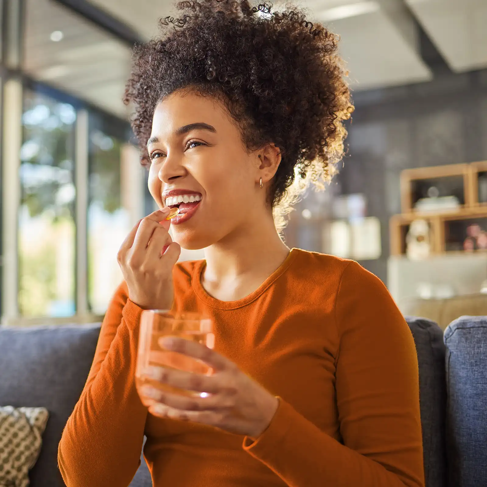 Eine Frau mit orangenem Oberteil nimmt Nahrungsergänzungsmittel. In der einen Hand hat sie die Kapsel und in der andern ein Glas Wasser. | VIACTIV Krankenkasse
