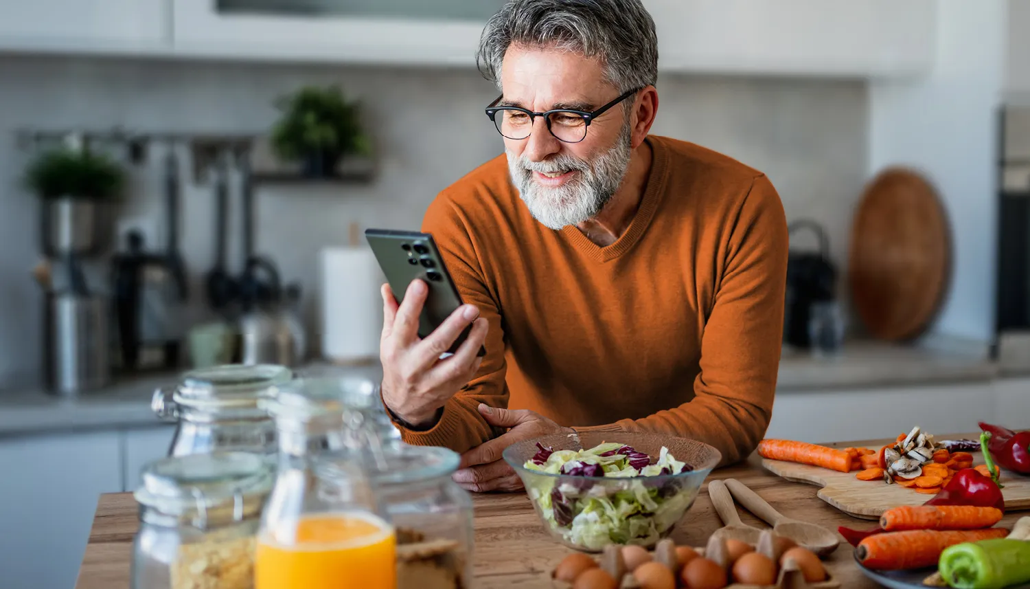 Ein Mann lehnt sich auf die Arbeitsfläche seiner Küche und führt ein Videogespräch über sein Smartphone. Neben ihm sieht man klein geschnittenes Gemüse. | VIACTIV Krankenkasse