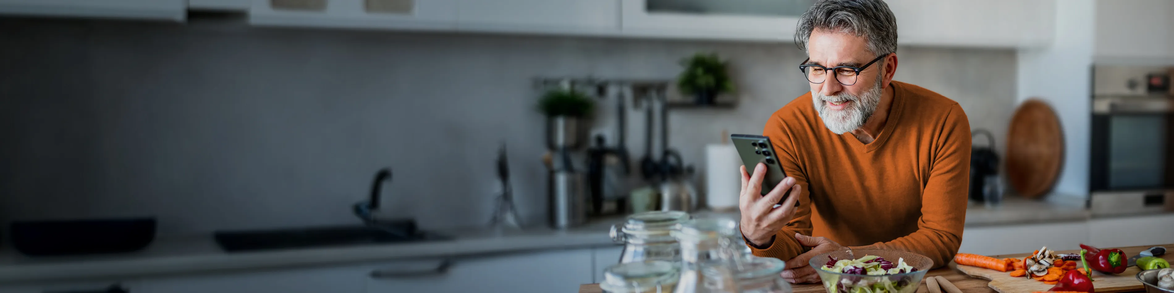 Ein Mann lehnt sich auf die Arbeitsfläche seiner Küche und führt ein Videogespräch über sein Smartphone. Neben ihm sieht man klein geschnittenes Gemüse. | VIACTIV Krankenkasse