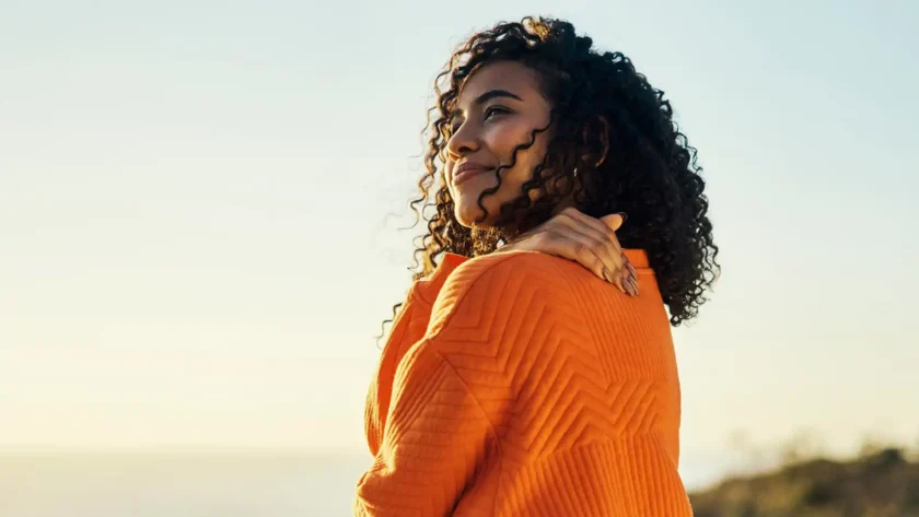 Eine Frau steht sehr entspannt draußen und lächelt. Mit der Hand greift sie sich an die Schulter. | VIACTIV Krankenkasse