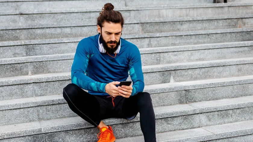 Ein Mann in Sportkleidung sitzt auf einer Treppe und schaut dabei auf sein Smartphone, welches er in der Hand hält. | VIACTIV Krankenkasse