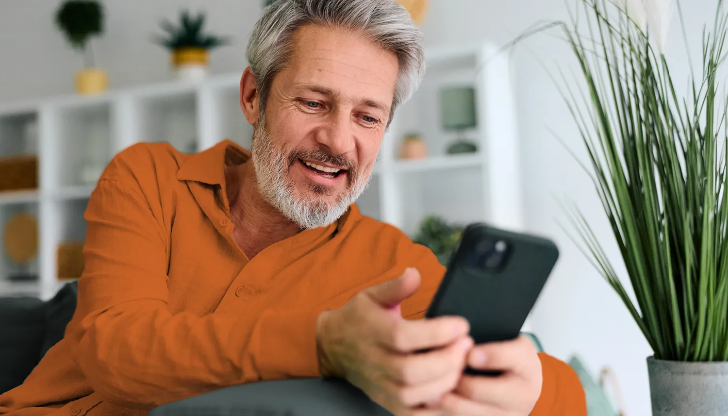 Ein Mann sitzt auf den Sofa und lächelt. Er hält ein Handy in der Hand und schaut sich die Manoa Plus App an. | VIACTIV Krankenkasse