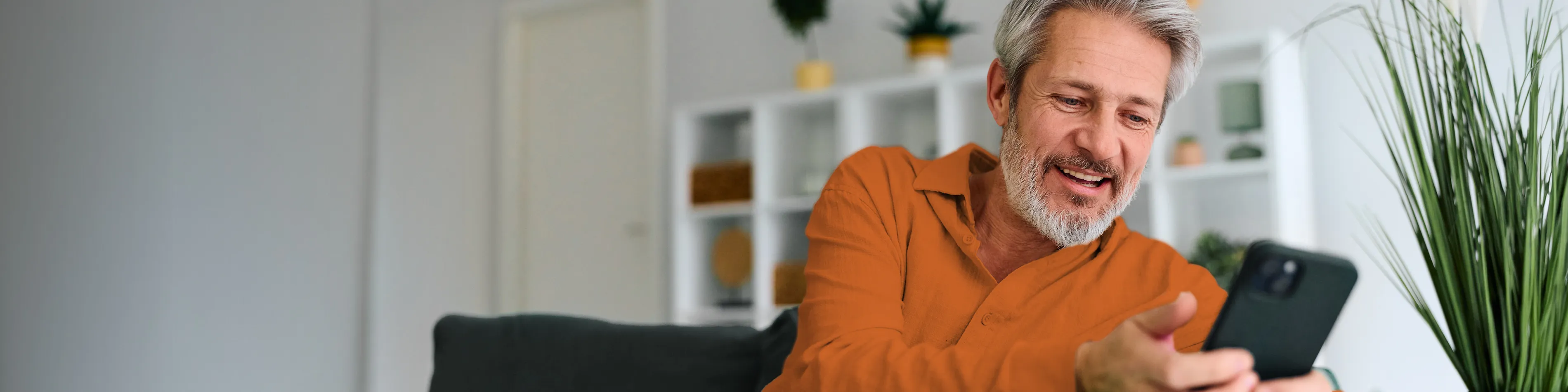 Ein Mann sitzt auf den Sofa und lächelt. Er hält ein Handy in der Hand und schaut sich die Manoa Plus App an. | VIACTIV Krankenkasse