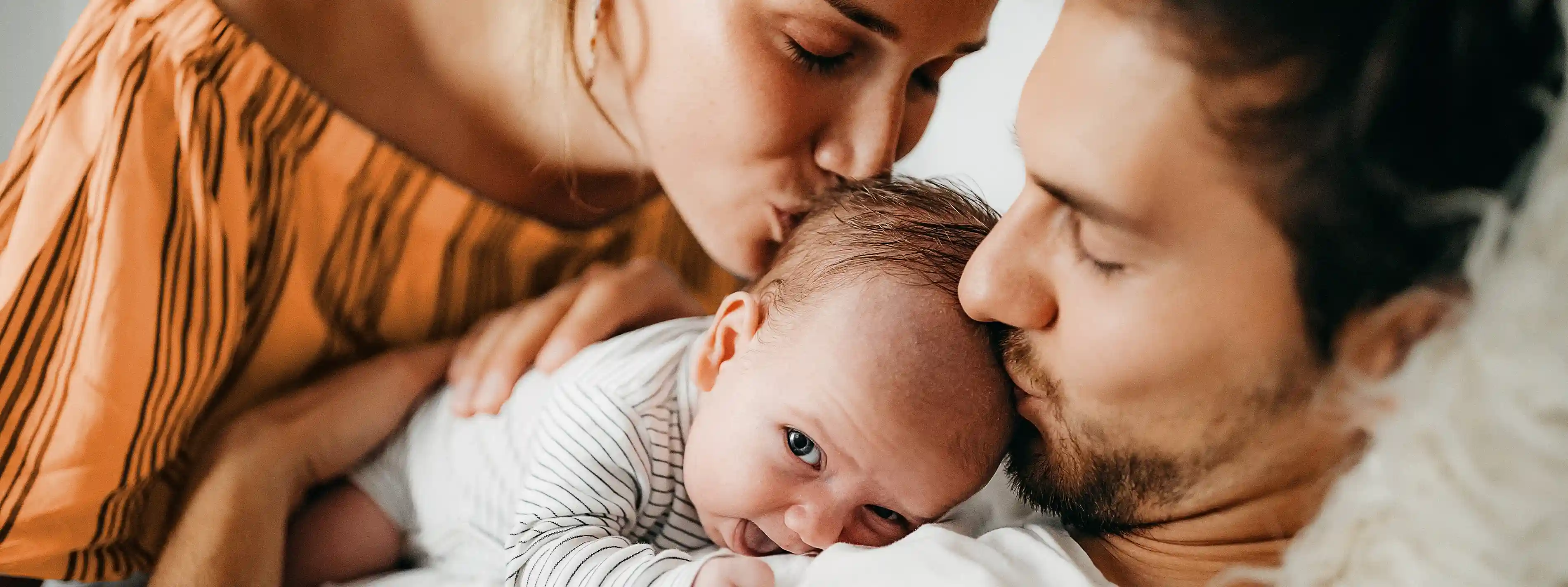 Eine Frau und ein Mann haben ein Baby in ihrer Mitte und beide küssen es auf den Kopf. | VIACTIV Krankenkasse