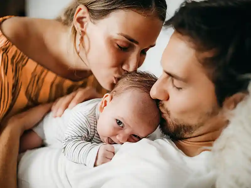 Eine Frau und ein Mann haben ein Baby in ihrer Mitte und beide küssen es auf den Kopf. | VIACTIV Krankenkasse