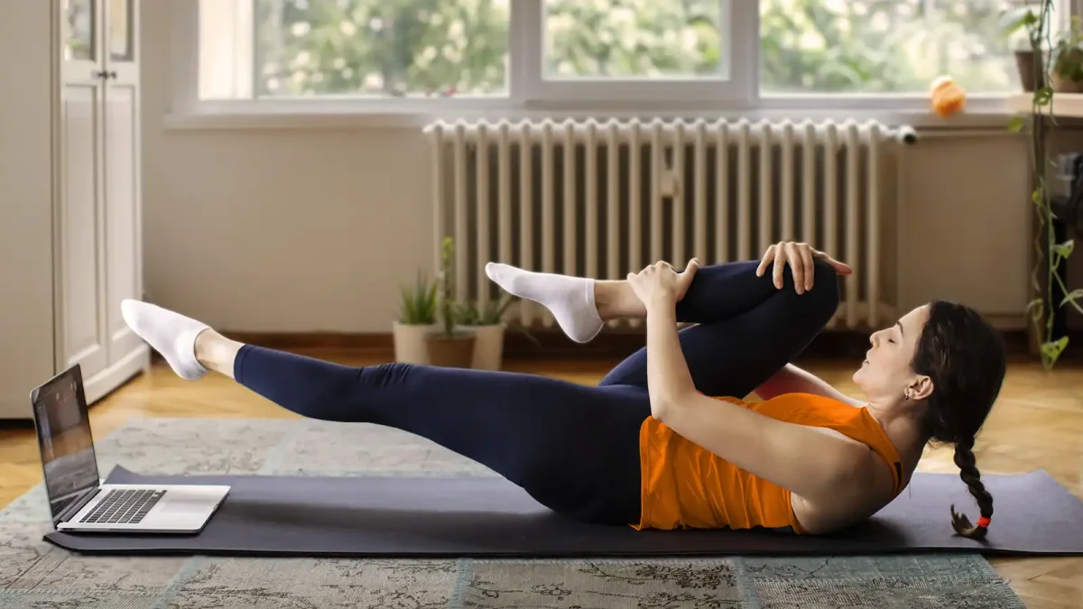 Eine Frau liegt in ihrem Wohnzimmer auf der Matte und macht eine Sportübung. Vor ihr steht ein Laptop, auf den sie blickt. | VIACTIV Krankenkasse