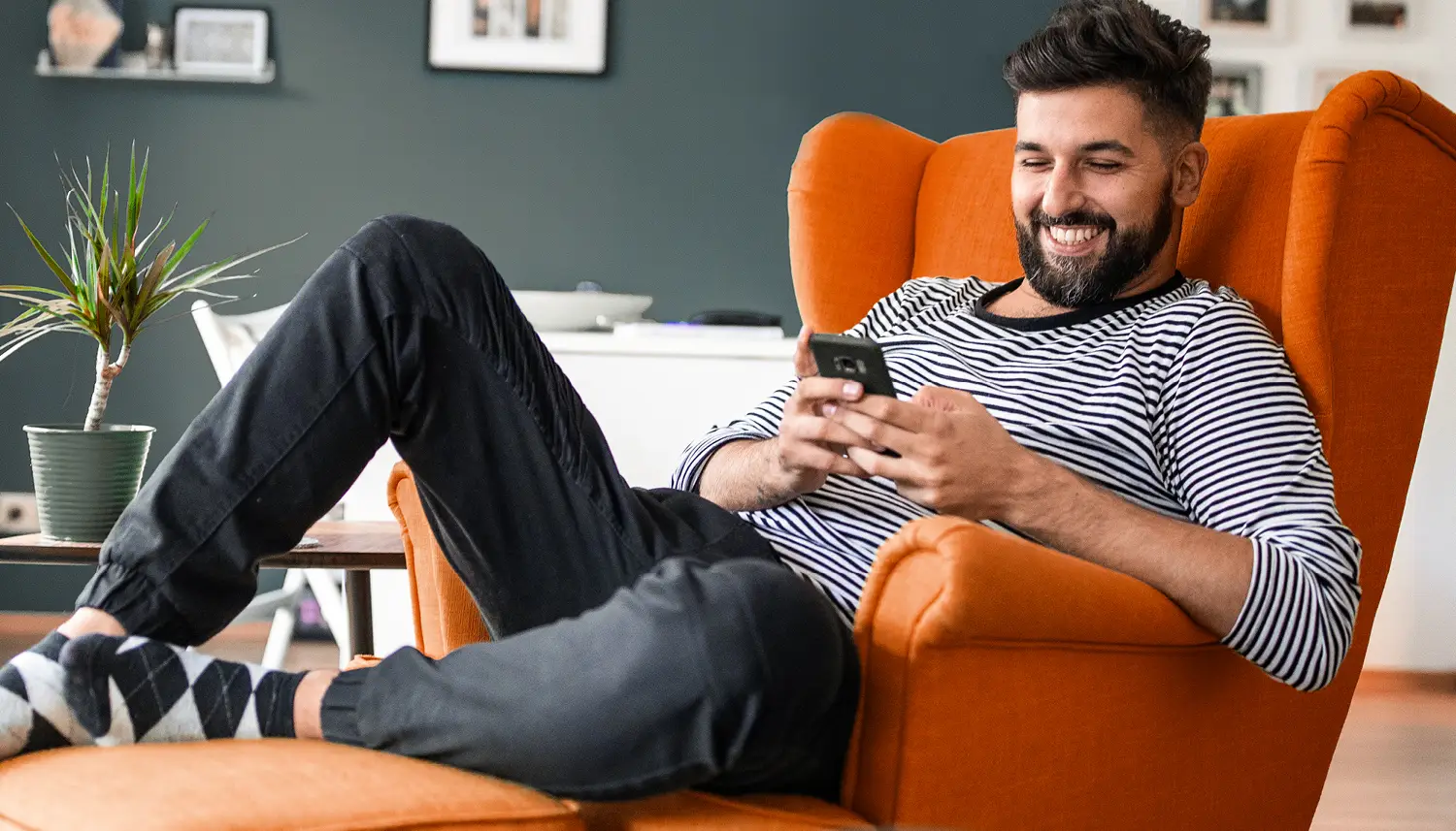 Ein junger Mann sitzt im Sessel und schaut amüsiert auf das Display seines Handys. | VIACTIV Krankenkasse