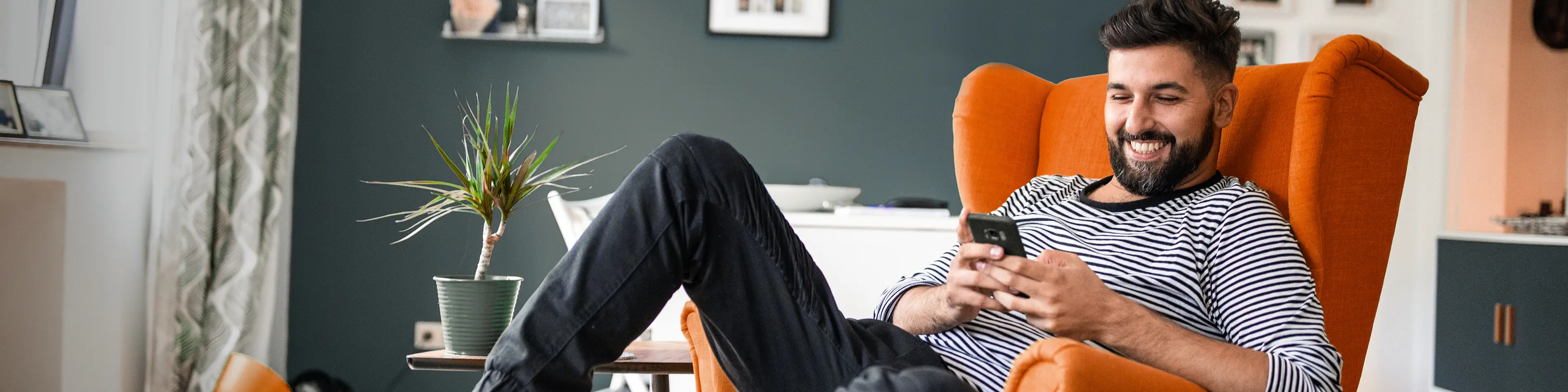 Ein junger Mann sitzt im Sessel und schaut amüsiert auf das Display seines Handys. | VIACTIV Krankenkasse