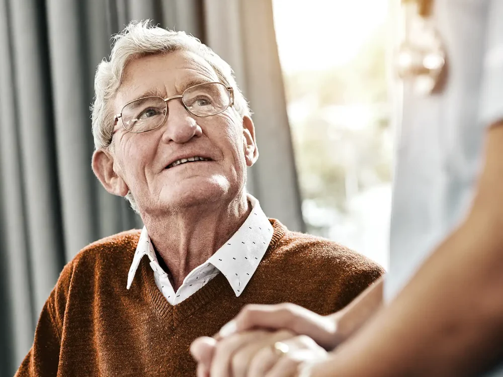 Ein älterer Mann blickt zur Pflegekraft auf und sie hält seine Hand. | VIACTIV Krankenkasse