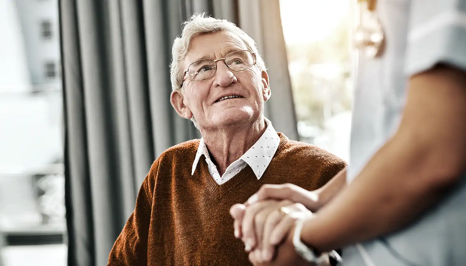 Ein älterer Mann blickt zur Pflegekraft auf und sie hält seine Hand. | VIACTIV Krankenkasse