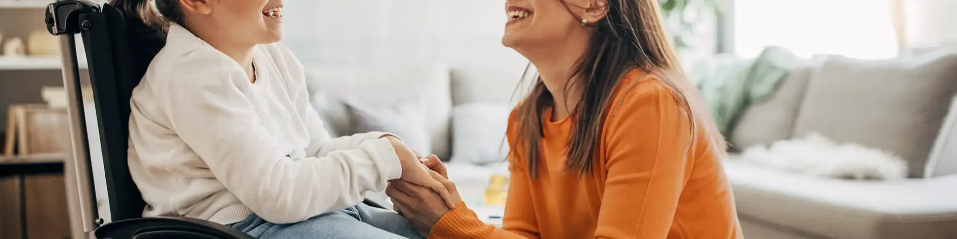 Eine junge Frau kniet vor einem Mädchen, die im Rollstuhl sitzt und hält ihre Hand. Sie lächeln. | VIACTIV Krankenkasse