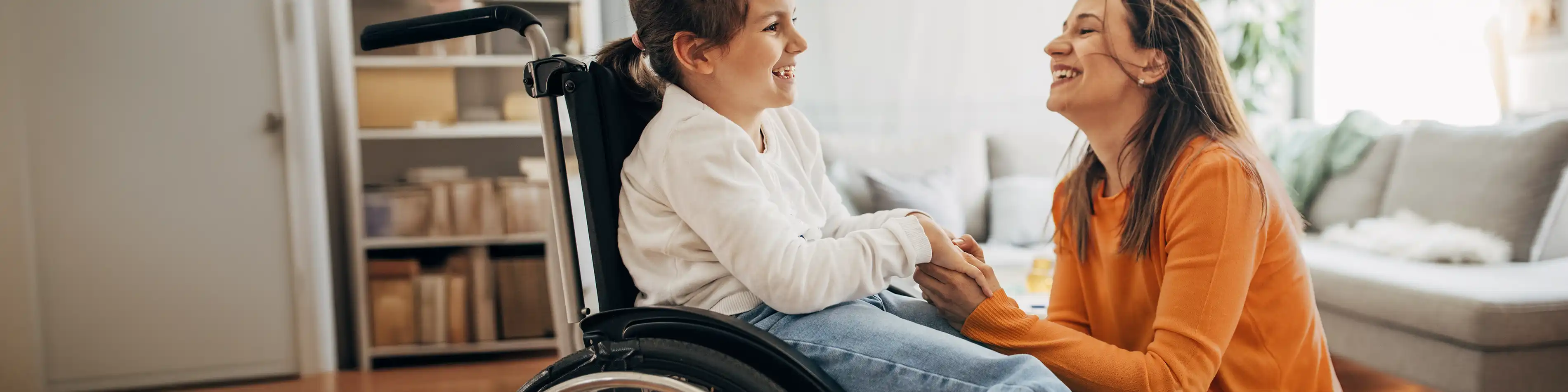 Eine junge Frau kniet vor einem Mädchen, die im Rollstuhl sitzt und hält ihre Hand. Sie lächeln. | VIACTIV Krankenkasse