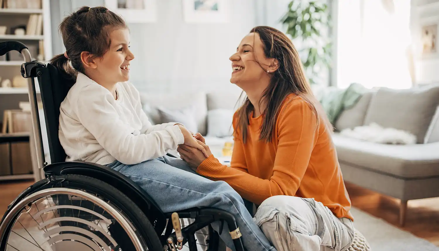 Eine junge Frau kniet vor einem Mädchen, die im Rollstuhl sitzt und hält ihre Hand. Sie lächeln. | VIACTIV Krankenkasse