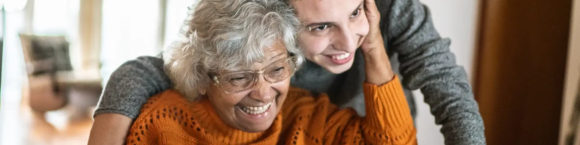 Eine ältere Frau sitzt vor dem Laptop. Ein junger Mann steht eng hinter ihr und sie legt die Hand auf seine Wange. | VIACTIV Krankenkasse