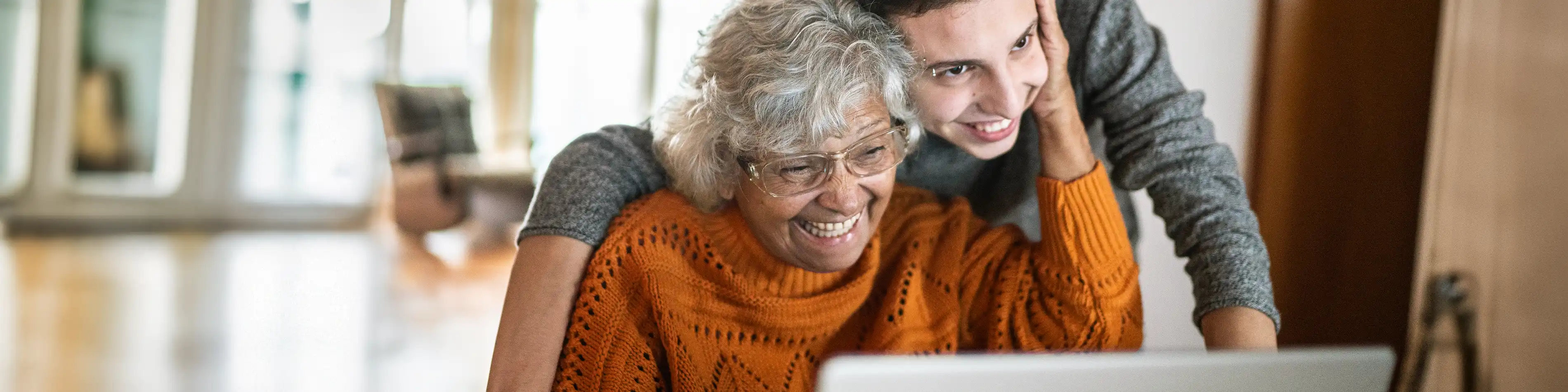 Eine ältere Frau sitzt vor dem Laptop. Ein junger Mann steht eng hinter ihr und sie legt die Hand auf seine Wange. | VIACTIV Krankenkasse