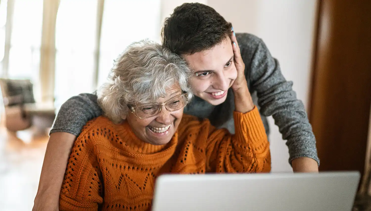 Eine ältere Frau sitzt vor dem Laptop. Ein junger Mann steht eng hinter ihr und sie legt die Hand auf seine Wange. | VIACTIV Krankenkasse