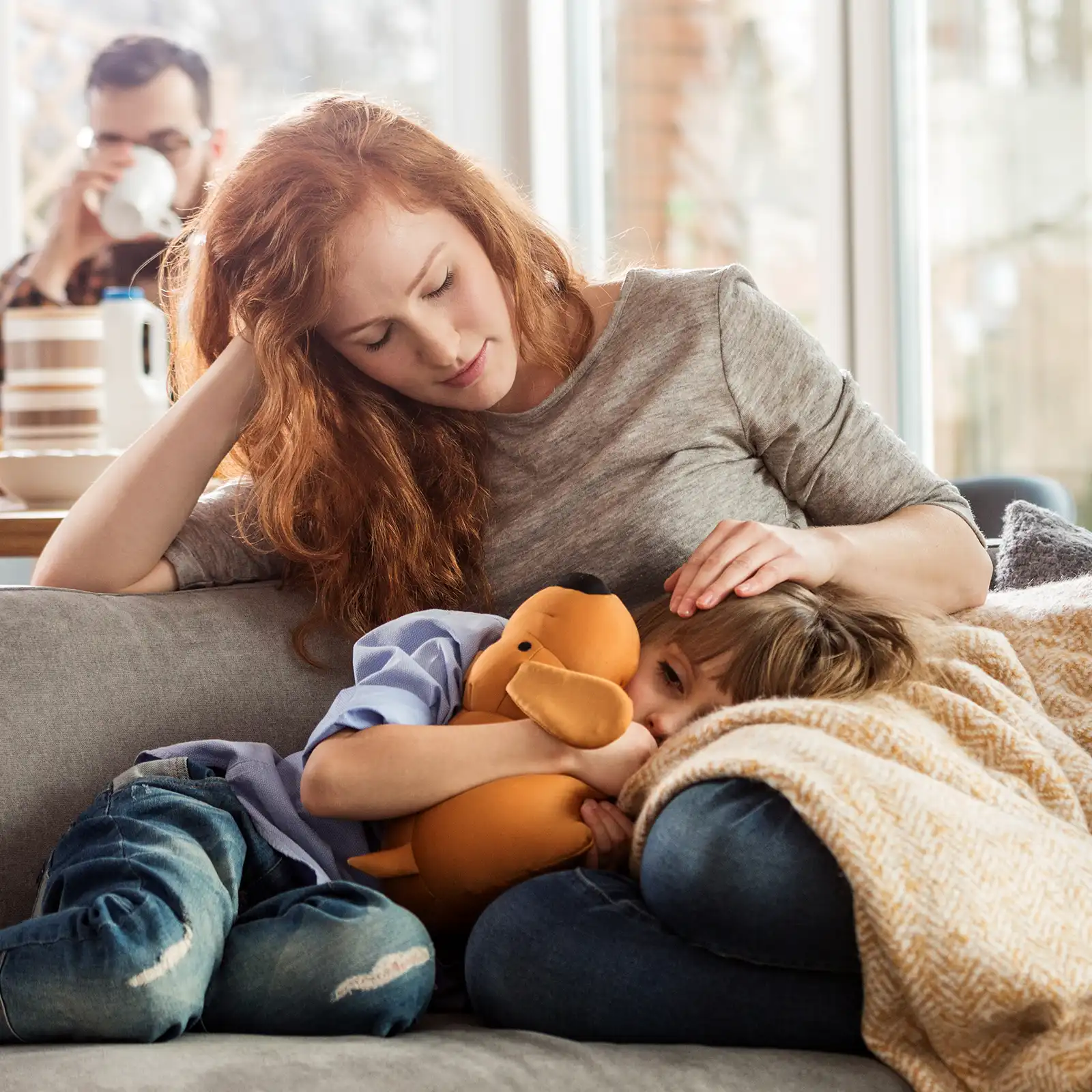 Eine junge Mutter sitzt auf dem Sofa. Ihr Sohn liegt mit dem Kopf auf ihrem Schoss. Der kleine Junge wirkt müde und krank und wird von der Mutter am Kopf gestreichelt. | VIACTIV Krankenkasse