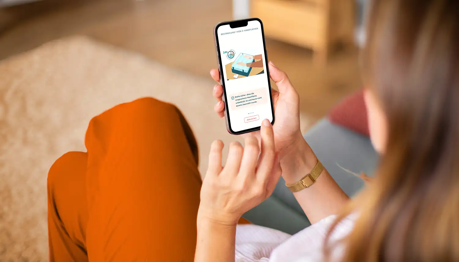 Eine Frau sitzt auf einem Sofa und schaut sich über ihr Smartphone die App Skinuvita an. | VIACTIV Krankenkasse