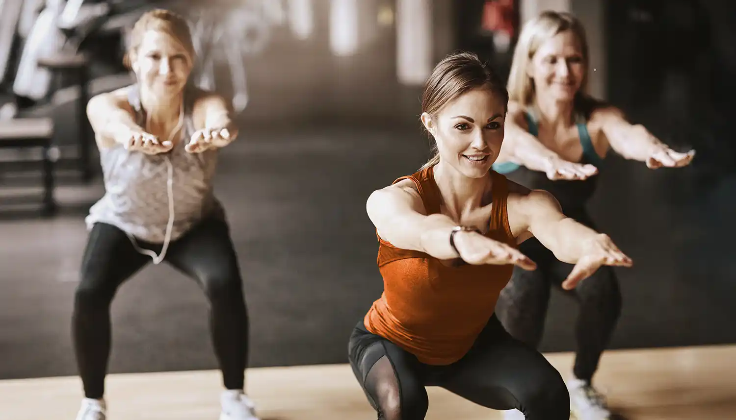 Eine Gruppe von jungen Frauen trainiert im Fitnessstudio und sie machen Kniebeugen. | VIACTIV Krankenkasse