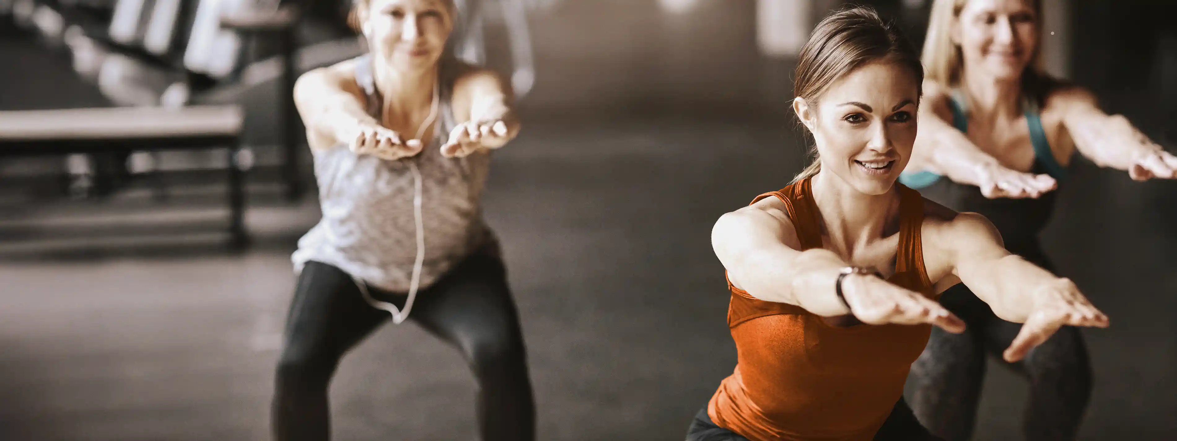 Eine Gruppe von jungen Frauen trainiert im Fitnessstudio und sie machen Kniebeugen. | VIACTIV Krankenkasse