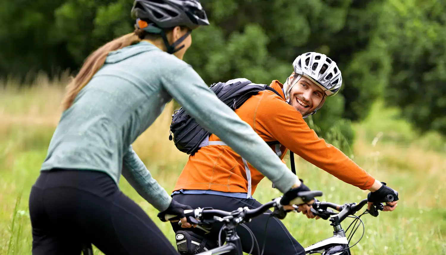 Zwei Fahrradfahrer sind in der Natur unterwegs. Der vordere Fahrer blickt über die Schulter zurück und lächelt. | VIACTIV Krankenkasse