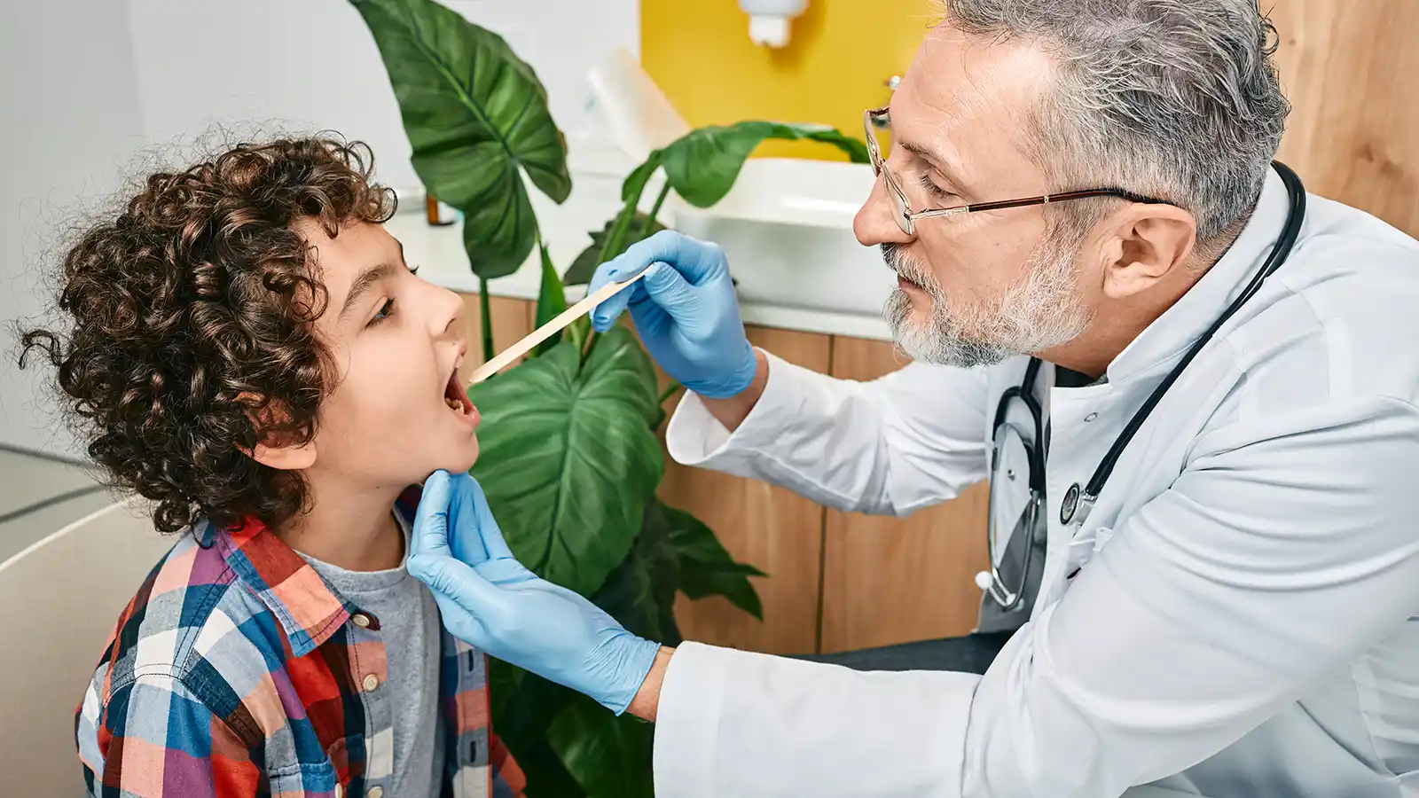 Ein Arzt schaut mithilfe einer Mundspatel in den Hals- und Mundraum eines Kindes. | VIACTIV Krankenkasse