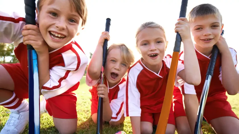 Eine Gruppe mit vier Kindern lächelt direkt in die Kamera. Sie halten Hockeyschläger in der Hand und tragen alle das gleiche Sportoutfit. | VIACTIV Krankenkasse
