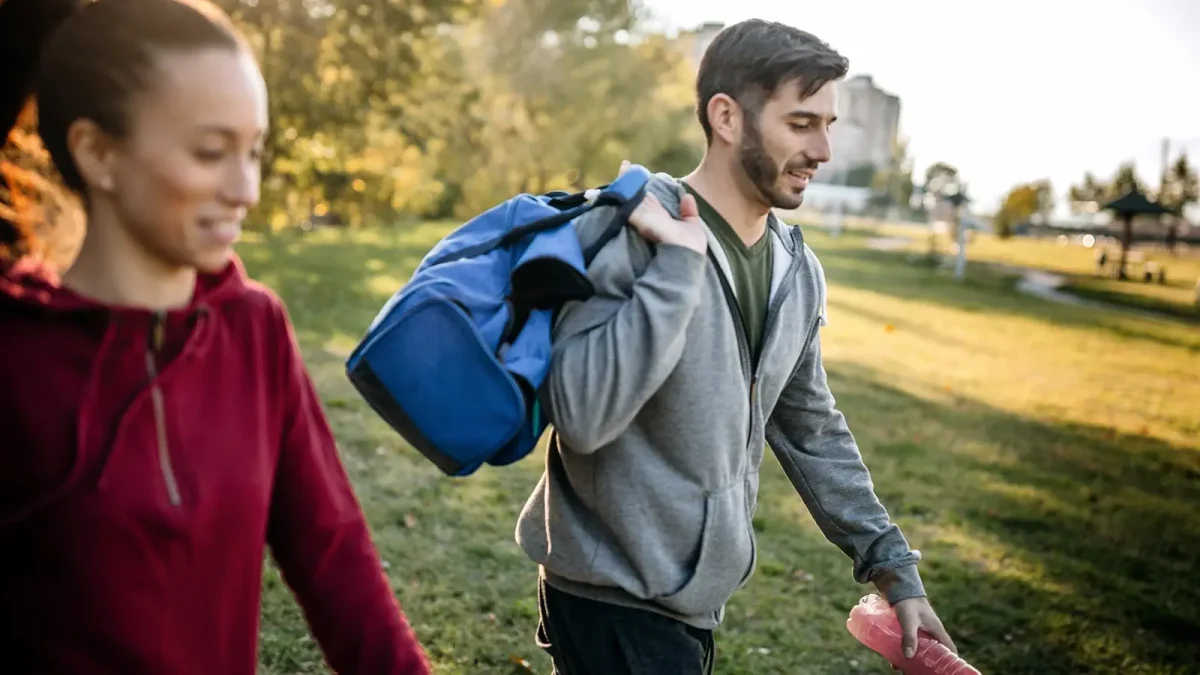 Junger Mann und junge Frau im Park bei Sonnenschein mit Sporttasche und Getränk auf dem Weg zum Outdoor Fitness. | VIACTIV Krankenkasse