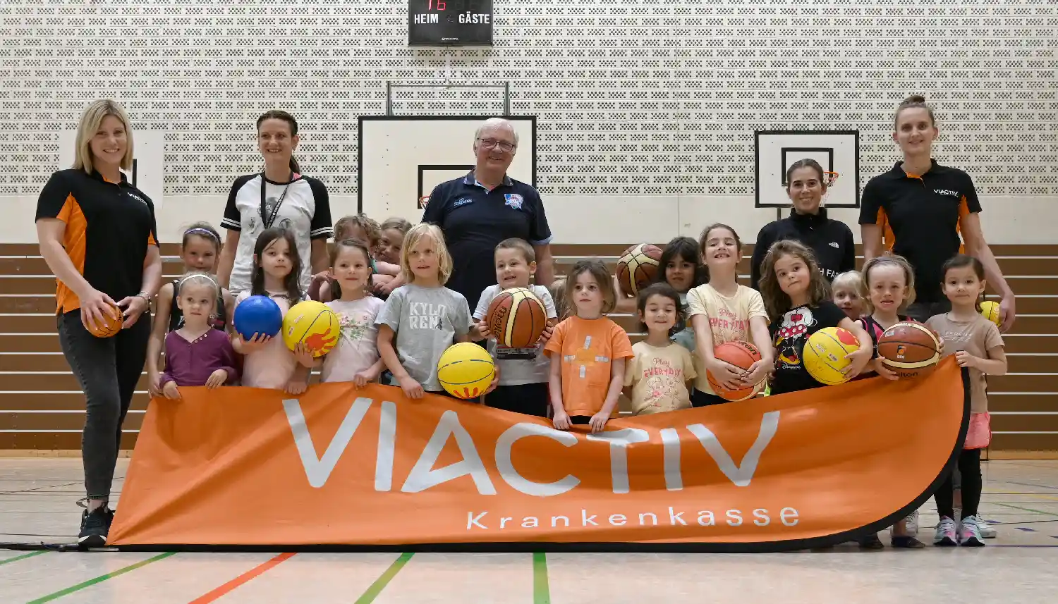 Das Gruppenfoto zeigt eine Gruppe kleiner Kinder in einer Sporthalle. Viele haben einen Basketball in der Hand und lachen zufrieden in die Kamera. Sie halten ein Banner der VIACTIV Krankenkasse hoch. Die VIACTIV organisiert die Aktion VIACTIV AstroKids. | VIACTIV Krankenkasse