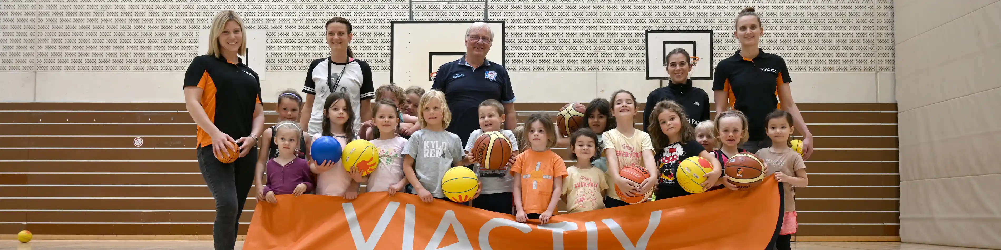 Das Gruppenfoto zeigt eine Gruppe kleiner Kinder in einer Sporthalle. Viele haben einen Basketball in der Hand und lachen zufrieden in die Kamera. Sie halten ein Banner der VIACTIV Krankenkasse hoch. Die VIACTIV organisiert die Aktion VIACTIV AstroKids. | VIACTIV Krankenkasse