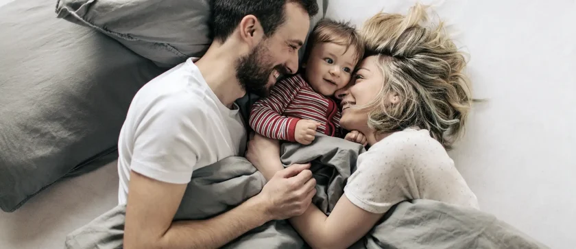 Eine Frau und ein Mann liegen sich zugewandt im Bett. Zwischen ihnen liegt ihr Kleinkind. Alle lächeln glücklich. | VIACTIV Krankenkasse